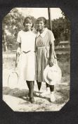 Two African American women stand outdoors posing beside a tennis racket in the 1920s. Both wear light-colored dresses and stylish accessories, smiling closely together in a relaxed portrait.