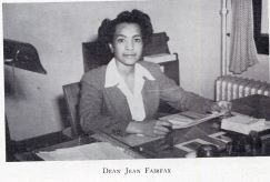 Black-and-white photograph of a woman identified as Dean Jean Fairfax seated at a desk in an office in 1945. She wears a tailored suit and looks toward the camera while surrounded by papers and office materials.
